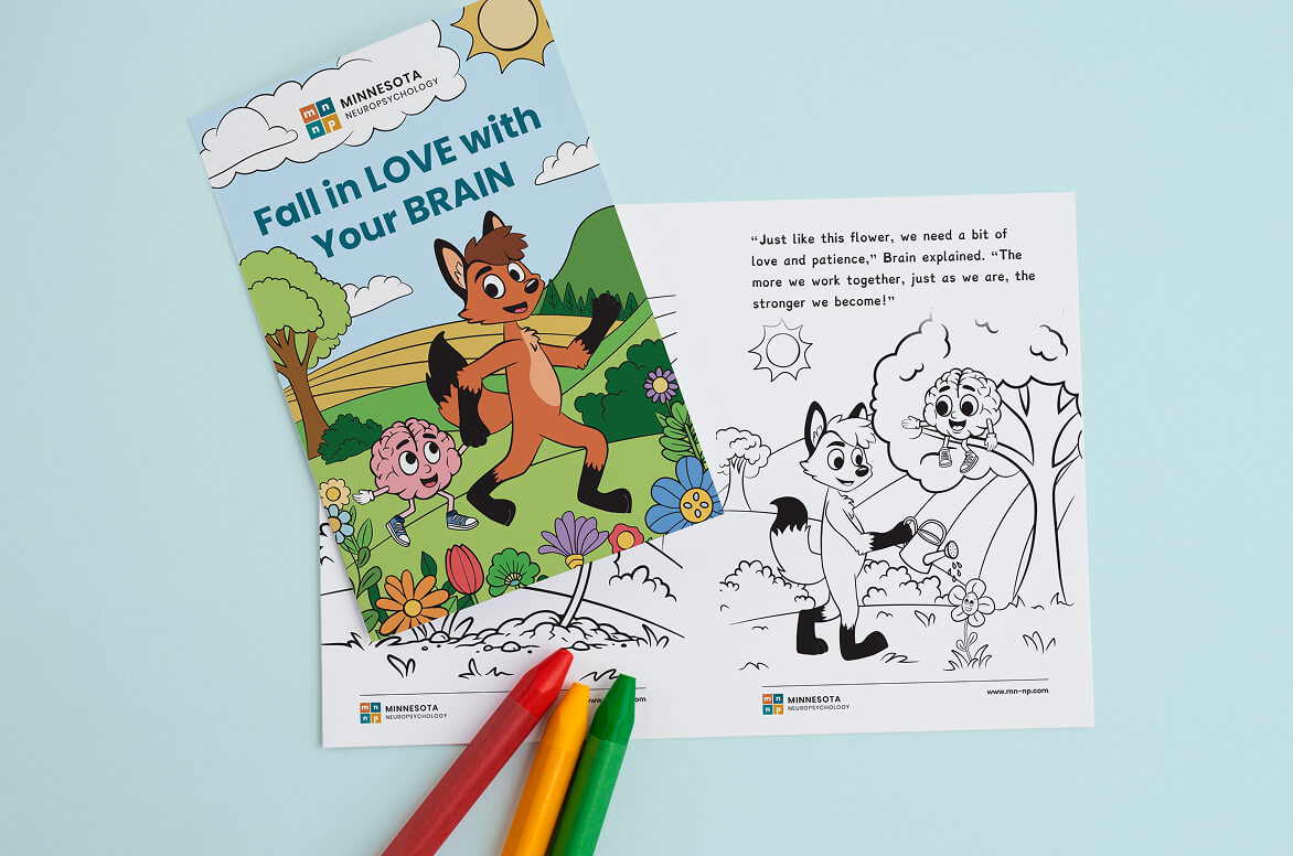 A children’s activity book titled “Fall in LOVE with Your BRAIN” lies open on a light blue surface. The colorful front cover shows a cartoon fox and a smiling brain character walking through a flower-filled landscape, while the open inside page features a black-and-white coloring illustration of the same characters watering a flower, with a short inspirational quote at the top. Three crayons—red, yellow, and green—are placed below the book.