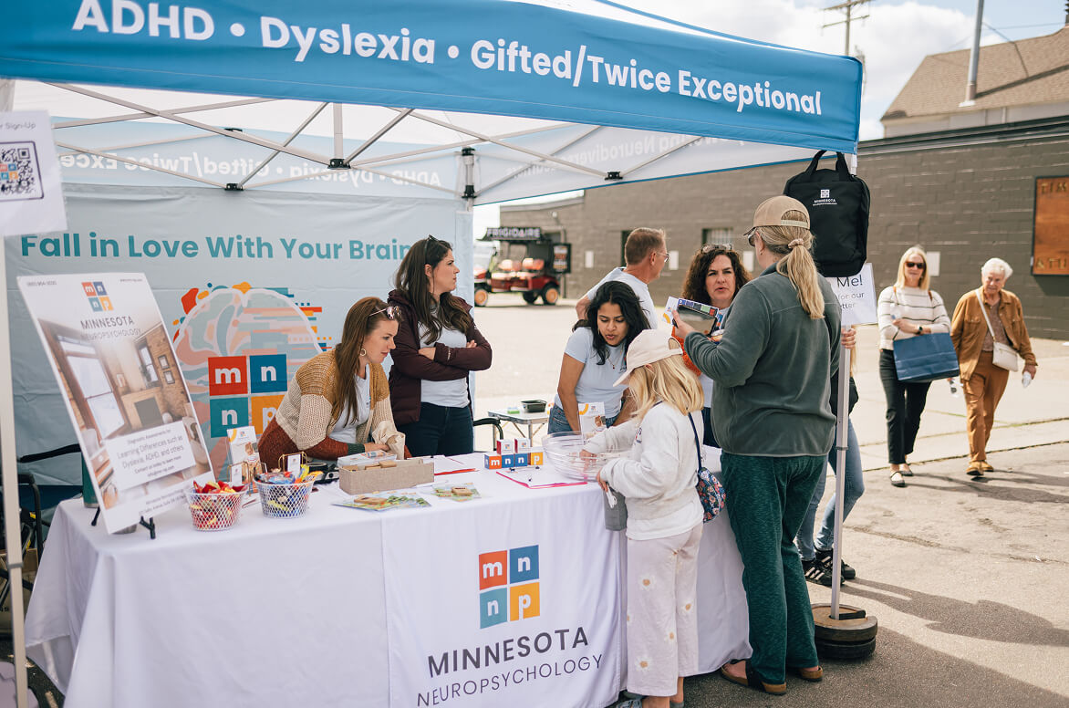 Community outreach booth for Minnesota Neuropsychology at outdoor event, with staff engaging visitors under tent displaying services for ADHD, dyslexia, and gifted individuals.