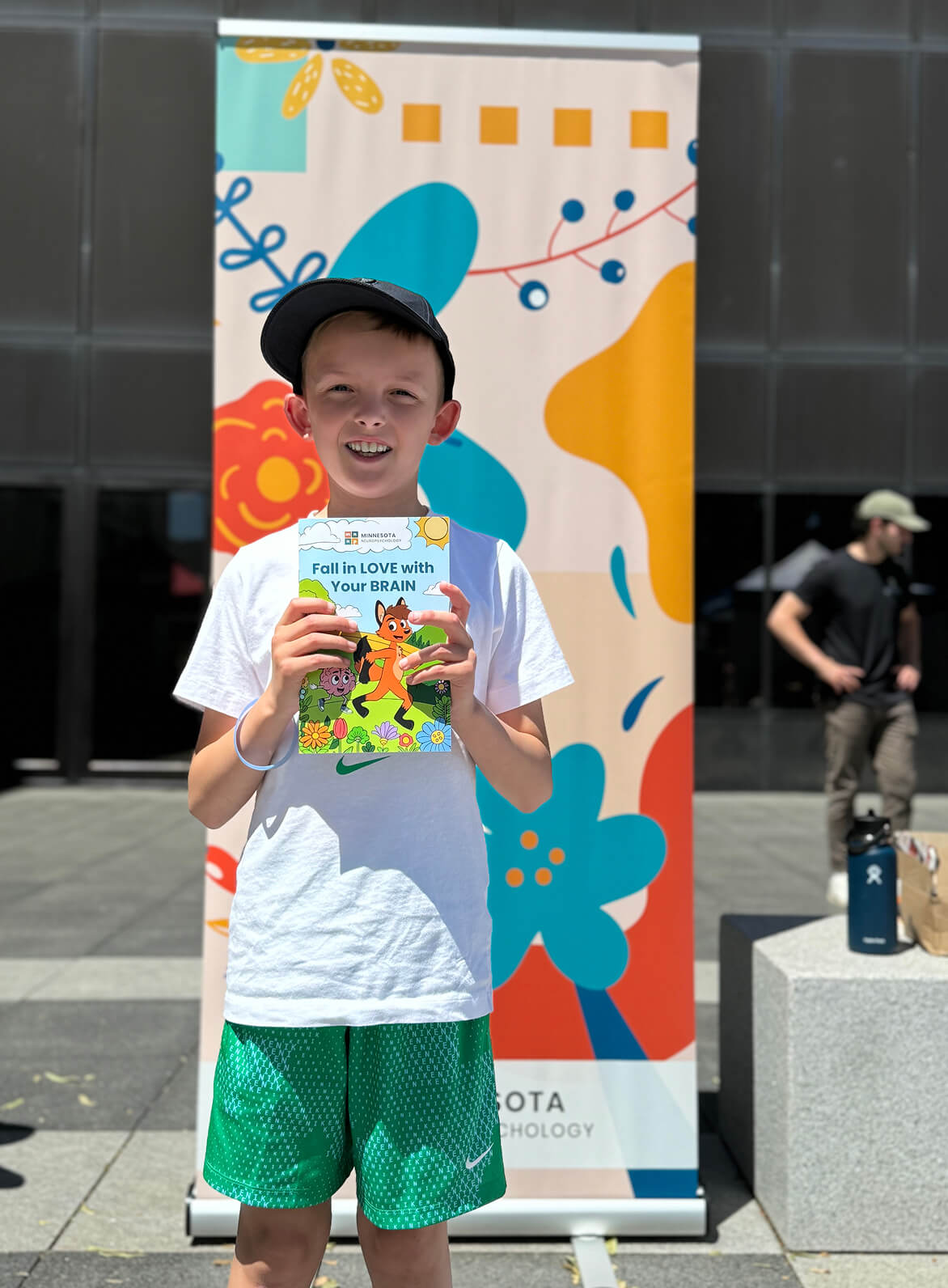 A smiling young child wearing a baseball cap, white T-shirt, and green shorts stands outdoors holding a colorful children’s book titled “Fall in LOVE with Your BRAIN.” Behind the child is a tall, bright banner with abstract shapes and flowers, and a modern building plaza is visible in the background.