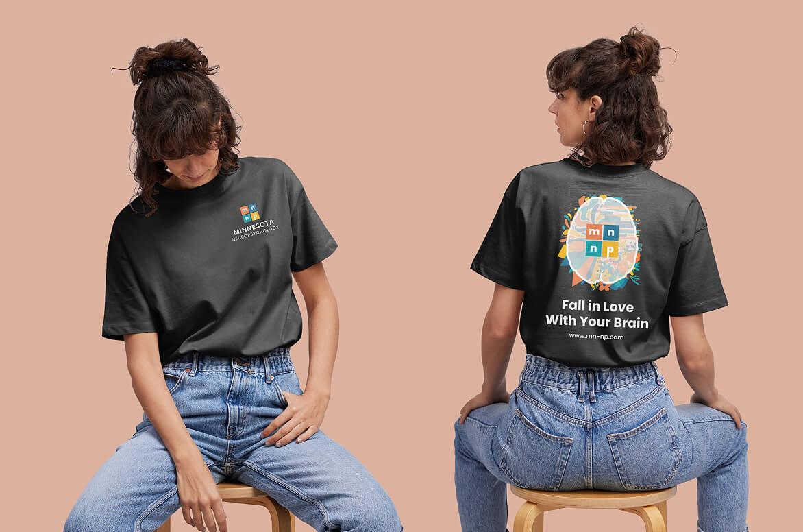 Front and back views of a person wearing a dark gray “Fall in Love With Your Brain” T-shirt with Minnesota Neuropsychology branding, paired with blue jeans, against a neutral background.